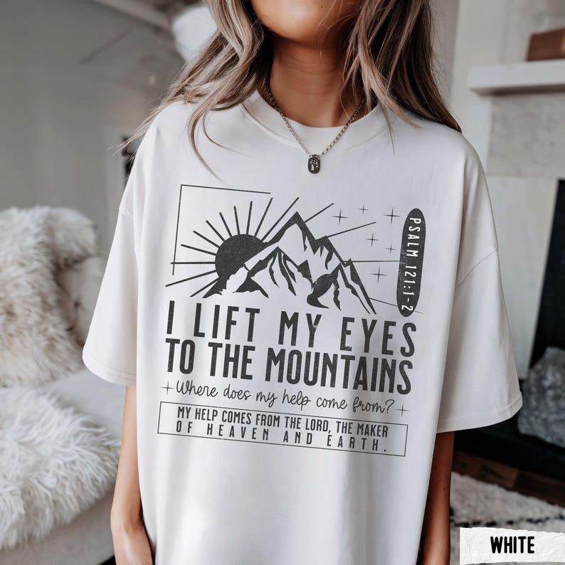 May include: White t-shirt with a black graphic of mountains, a rising sun, and the text "I Lift My Eyes to the Mountains." The shirt also includes the Bible verse Psalm 121:1-2 and the text "Where does my help come from?"