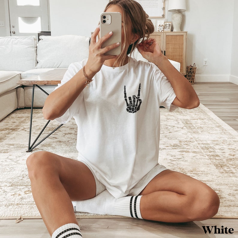 May include: White oversized t-shirt with a black skeleton hand making a rock and roll sign. The shirt is worn by a person sitting on a rug.
