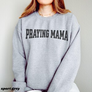 Pode incluir: Uma camisa de moletom cinza com o texto "Praying Mama" impresso em preto.