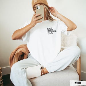Puede incluir: Camiseta blanca con el texto "Late Night Reading Club" impreso en la parte delantera. La modelo lleva la camiseta con un pantal&oacute;n de ch&aacute;ndal blanco.