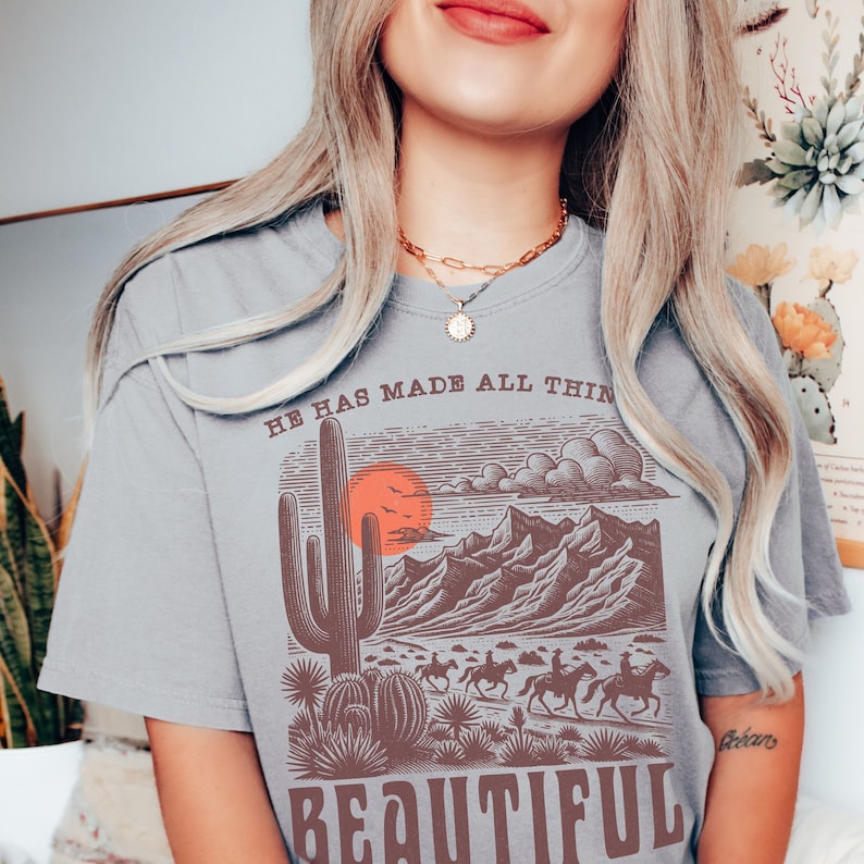 May include: Gray t-shirt with a graphic print of a desert landscape with a cactus, mountains, and a sunset. The text "He has made all things beautiful" is printed on the shirt.