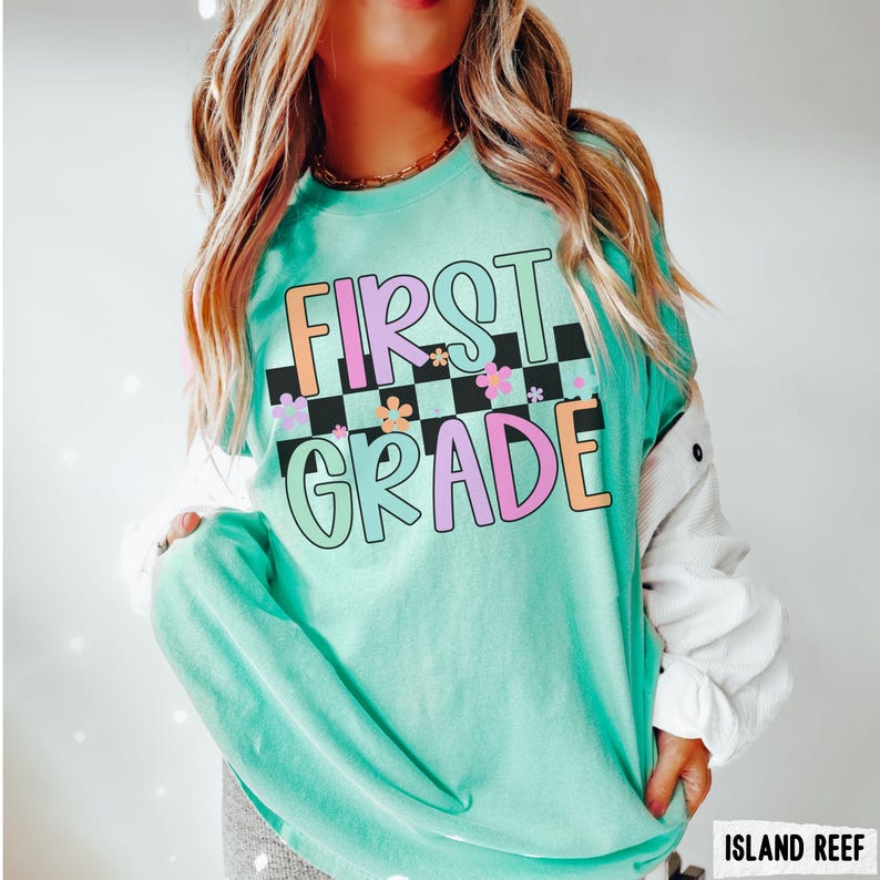 K&ouml;nnte beinhalten: Ein hellt&uuml;rkisfarbenes Langarmshirt mit dem Schriftzug "FIRST GRADE" in einer farbenfrohen, verspielten Schriftart. Der Text ist vor einem schwarz-wei&szlig; karierten Hintergrund mit floralen Akzenten platziert. Das Shirt ist ein bequemer, l&auml;ssiger Stil.