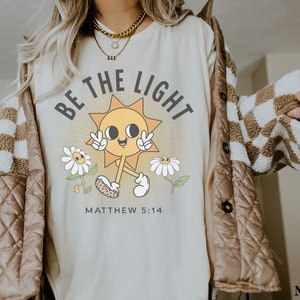 Op de afbeelding: Een beige t-shirt met een afbeelding van een lachende zon met de tekst "Be the Light" en "Matthew 5:14" eronder. De zon draagt ​​een vredesteken en is omringd door madeliefjes.