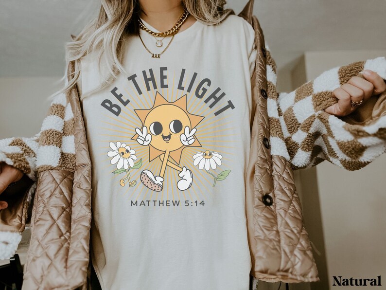 Op de afbeelding: Een beige t-shirt met een afbeelding van een lachende zon met de tekst "Be the Light" en "Matthew 5:14" eronder. De zon draagt ​​een vredesteken en is omringd door madeliefjes.