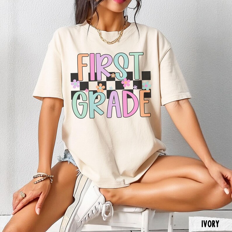 K&ouml;nnte beinhalten: Elfenbeinfarbenes T-Shirt mit den Worten "FIRST GRADE" in bunten, umrandeten Buchstaben. Der Text ist vor einem schwarz-wei&szlig; karierten Hintergrund mit Blumenakzenten platziert. Das Shirt ist ein l&auml;ssiger Stil mit kurzen &Auml;rmeln.