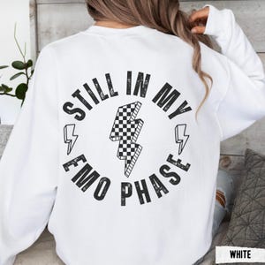 Pu&ograve; includere: Felpa bianca con un fulmine a scacchi in bianco e nero. Il testo sul retro recita "STILL IN MY EMO PHASE" in un design circolare con accenti di fulmini. La felpa &egrave; uno stile casual.