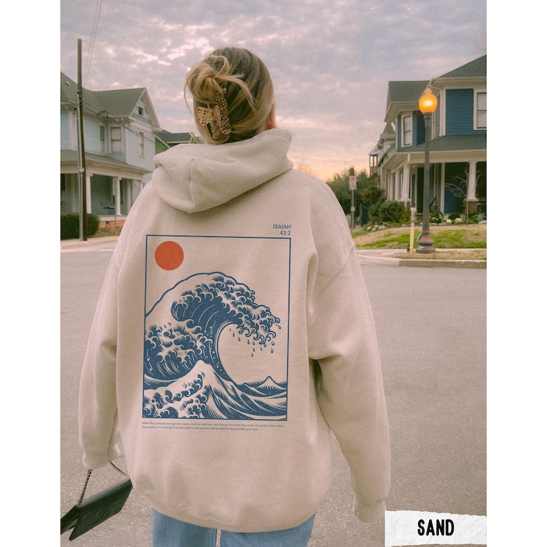 May include: A sand-colored hoodie with a large graphic on the back featuring a blue wave, a red sun, and the text "ISAIAH 43:2". The hoodie is being worn outdoors.