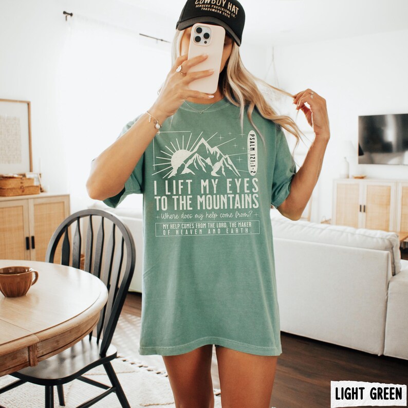 May include: Light green t-shirt with the text "I Lift My Eyes to the Mountains" and a mountain graphic. The shirt also includes the text "My help comes from the Lord, the maker of heaven and earth." The shirt is a light green color.