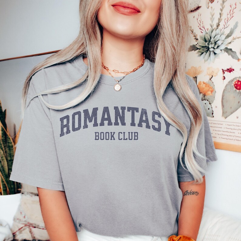 May include: A gray t-shirt with the text "ROMANTASY BOOK CLUB" printed in a navy blue collegiate font.