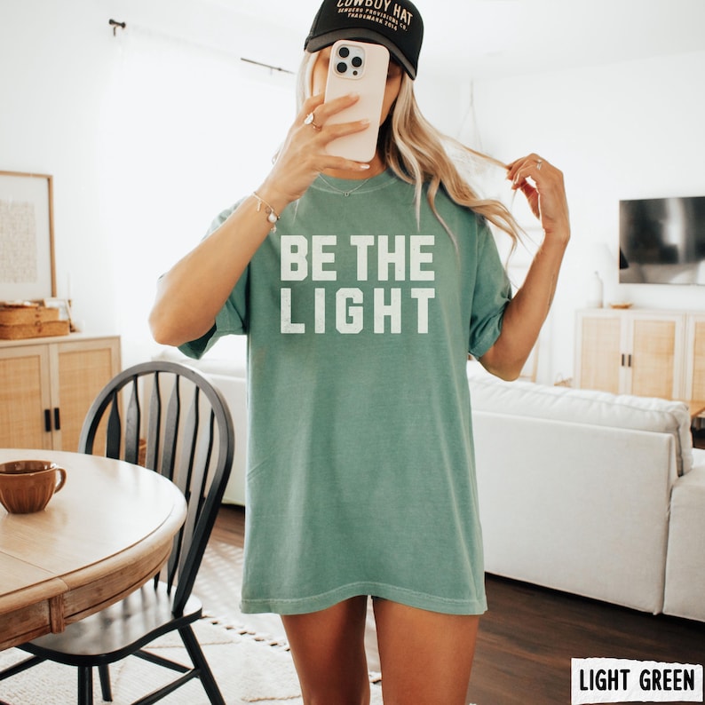 Peut inclure: T-shirt vert clair avec le texte "BE THE LIGHT" en lettres blanches. Le mod&egrave;le porte une casquette de baseball noire. Le t-shirt est de couleur vert clair.