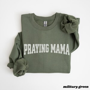 Pode incluir: Uma camisola verde com o texto "Praying Mama" em letras brancas. A camisola &eacute; de gola redonda e tem um caimento descontra&iacute;do.