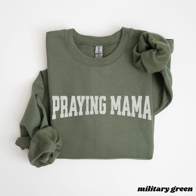 Pode incluir: Uma camisola verde com o texto "Praying Mama" em letras brancas. A camisola &eacute; de gola redonda e tem um caimento descontra&iacute;do.