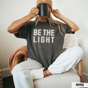 Peut inclure: T-shirt gris fonc&eacute; avec le texte "BE THE LIGHT" en lettres blanches. La personne porte un bonnet marron et tient une tasse noire. La personne porte un pantalon de surv&ecirc;tement gris clair et est assise sur une chaise.