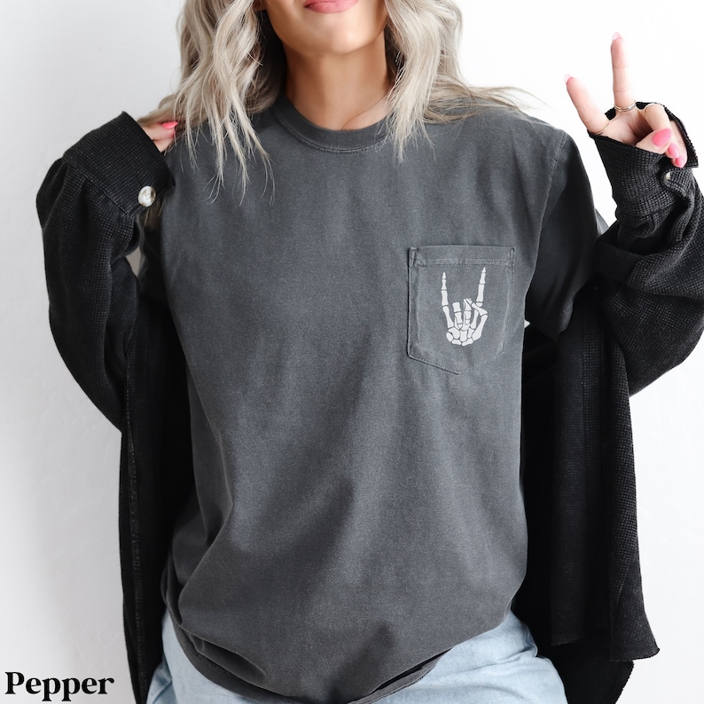 May include: A dark gray long-sleeved t-shirt with a pocket on the left side. The pocket has a white skeleton hand making a rock and roll sign.
