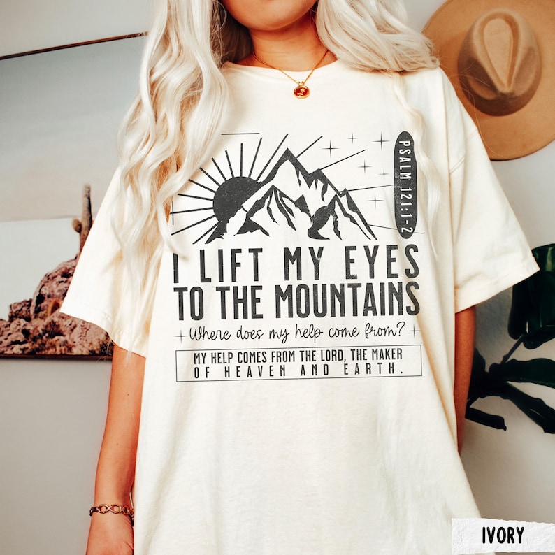 May include: Ivory-colored t-shirt with a mountain and sun graphic. The text reads "I Lift My Eyes To The Mountains" and "My Help Comes From The Lord, The Maker Of Heaven And Earth." Psalm 121:1-2 is also printed on the shirt.