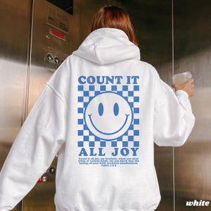 May include: White hoodie with a blue and white checkered smiley face graphic on the back. The text "COUNT IT ALL JOY" is above and below the smiley face. The hoodie also has text from James 1:2-3.