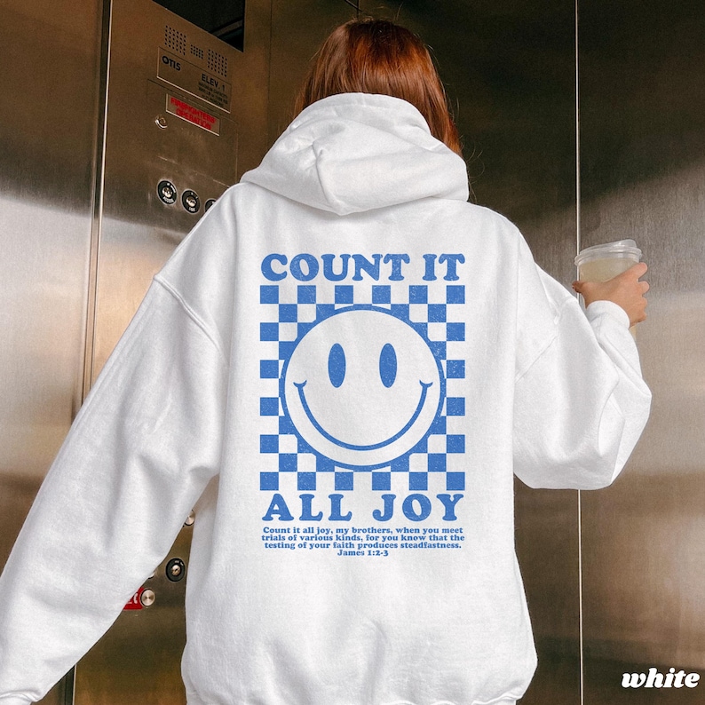 May include: White hoodie with a blue and white checkered smiley face graphic on the back. The text "COUNT IT ALL JOY" is above and below the smiley face. The hoodie also has text from James 1:2-3.