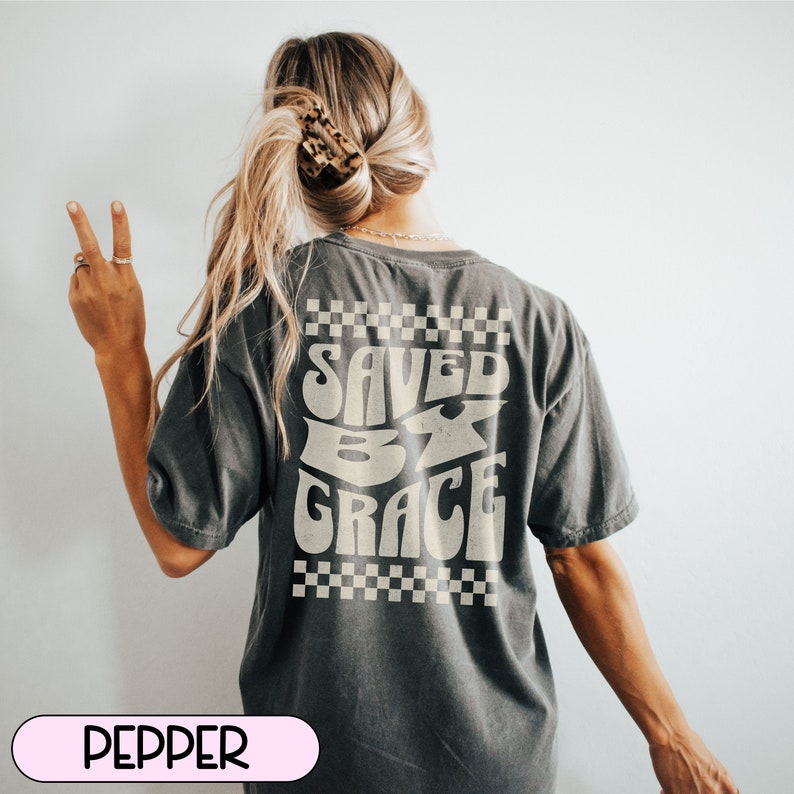 May include: A dark gray t-shirt with a white graphic that says "SAVED BY GRACE" in a retro font. The graphic is surrounded by a checkered pattern.