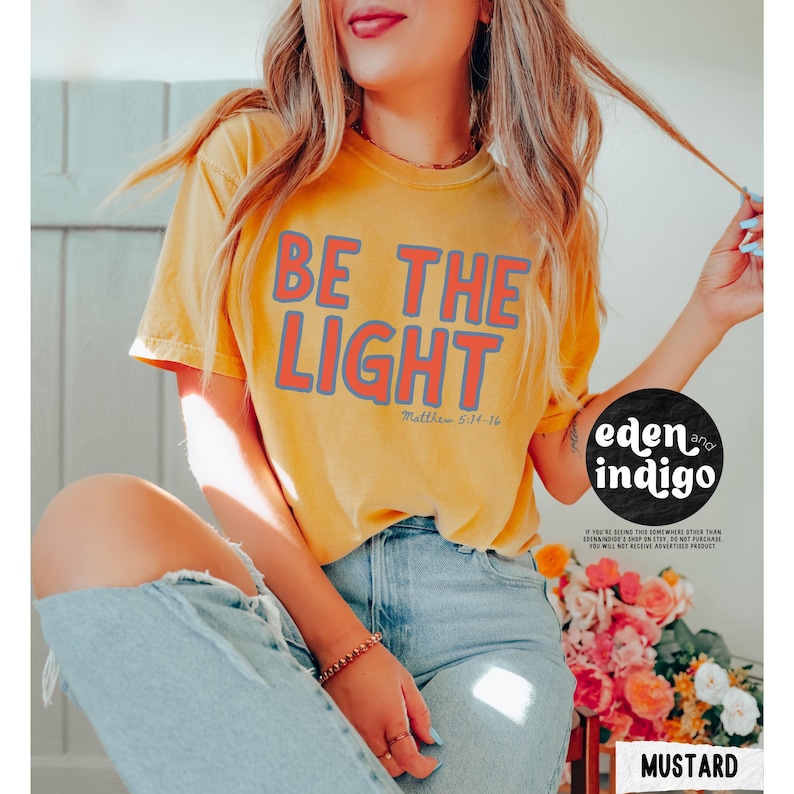 May include: Mustard yellow t-shirt with the text "BE THE LIGHT" in red and gray. The text "Matthew 5:14-16" is below the main text. The shirt is part of the "eden and indigo" collection.