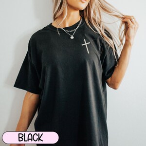 May include: A black oversized t-shirt with a small silver cross on the front. The shirt is being worn by a person with long blonde hair.