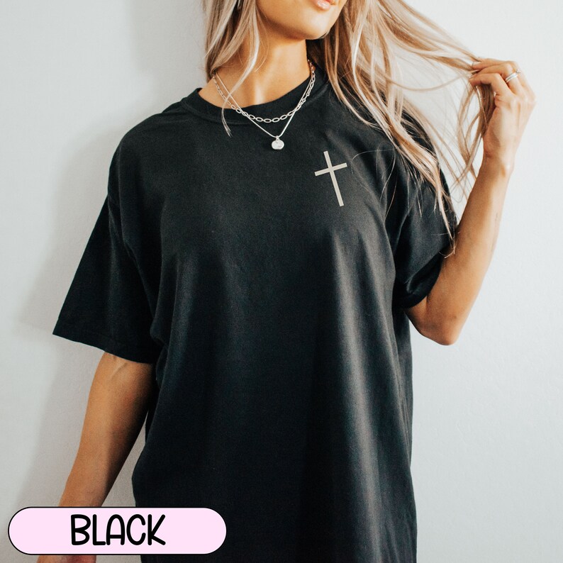 May include: A black oversized t-shirt with a small silver cross on the front. The shirt is being worn by a person with long blonde hair.