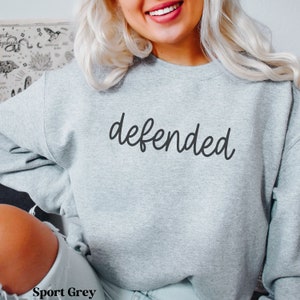May include: Sport Grey sweatshirt with the word "defended" in black script. The sweatshirt is a light gray color and has a crew neck. The text "Sport Grey" is printed at the bottom of the image.