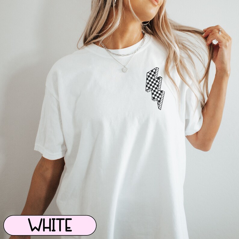 May include: White t-shirt with a black and white checkered lightning bolt graphic on the front. The word "WHITE" is displayed in a pink oval at the bottom of the image. The shirt has short sleeves and a relaxed fit.