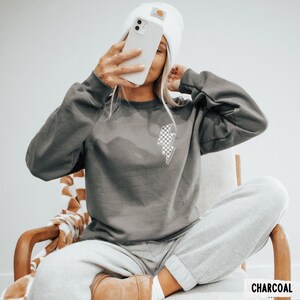 Pu&ograve; includere: Una felpa grigio antracite con una grafica a fulmine a scacchi. La felpa &egrave; abbinata a pantaloni della tuta grigi e un berretto bianco. La parola "CHARCOAL" &egrave; stampata in bianco in basso a destra nell'immagine.