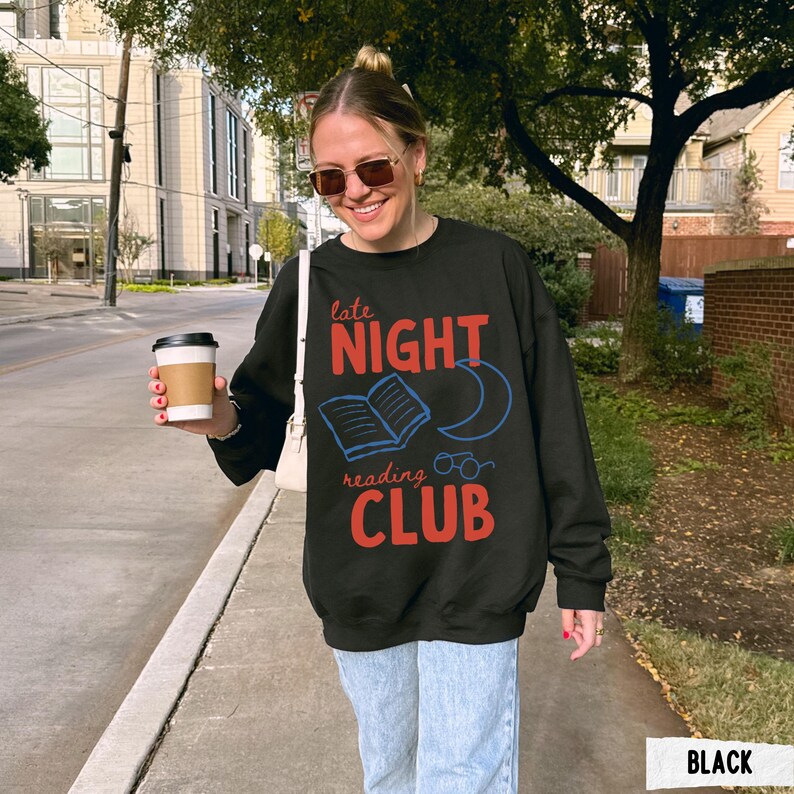 May include: Black sweatshirt with the text "late NIGHT reading CLUB" in red and blue. The design includes an open book, a crescent moon, and glasses. The sweatshirt is worn by a person holding a coffee cup.