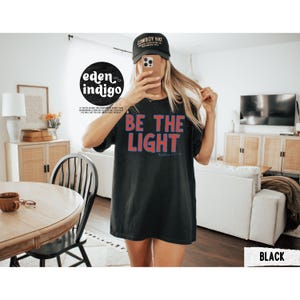 May include: Black t-shirt with the words "BE THE LIGHT" in red and blue lettering. The text "Matthew 5:14-16" is below the main text. The shirt is part of a clothing collection.