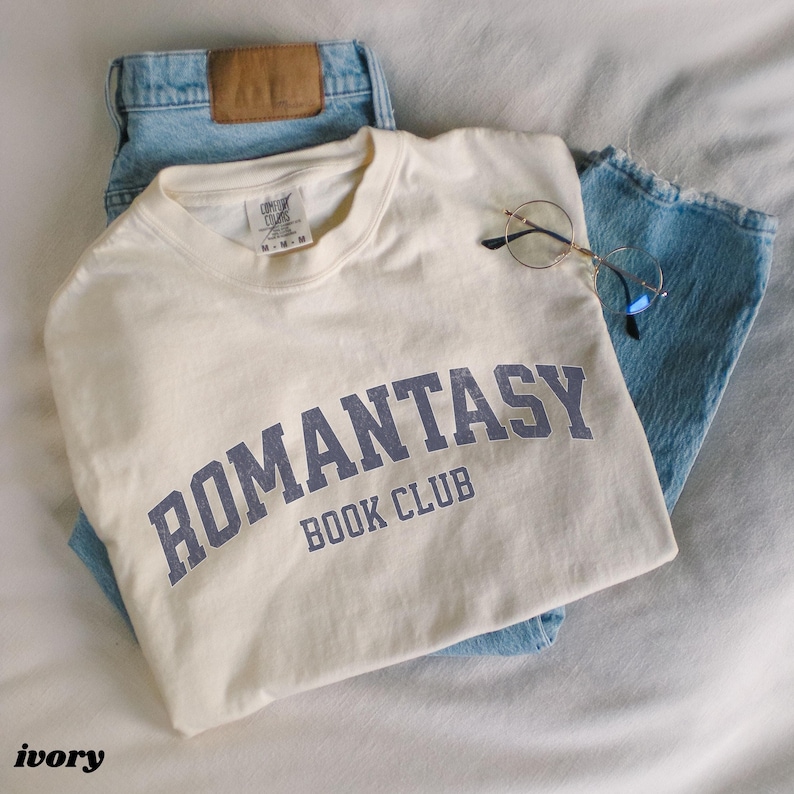 May include: A white t-shirt with the text "ROMANTASY BOOK CLUB" in blue lettering. The shirt is folded and placed on top of a pair of blue jeans.