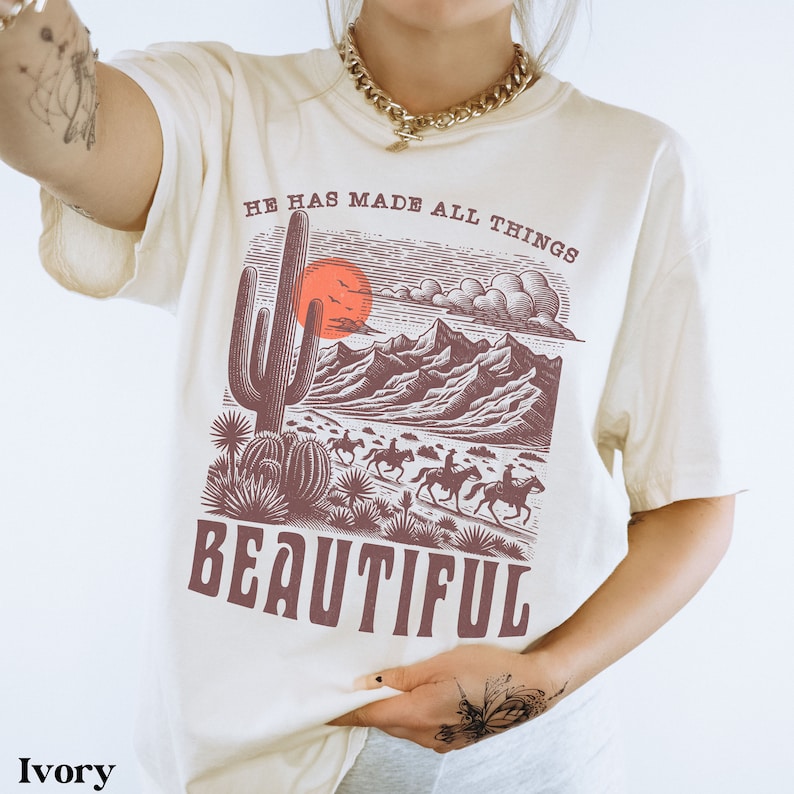 K&ouml;nnte beinhalten: Ein T-Shirt in Elfenbein mit einem grafischen Druck einer W&uuml;stenlandschaft mit einem Kaktus, Bergen und einem Sonnenuntergang. Der Text "He has made all things beautiful" ist auf das T-Shirt gedruckt.