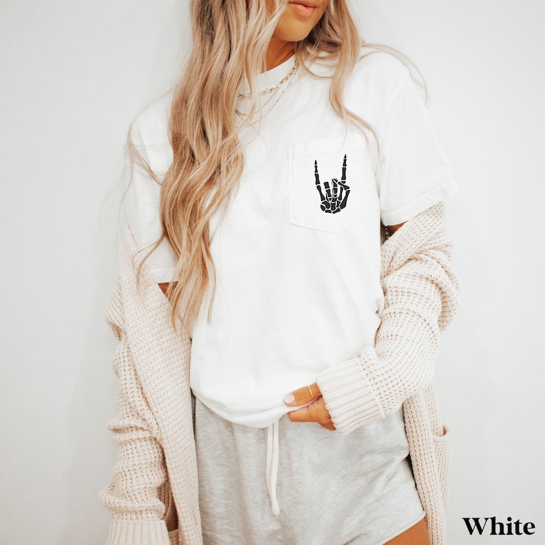 May include: White t-shirt with a black skeleton hand making a rock and roll sign in a pocket on the front of the shirt. The shirt is being worn by a person with long blonde hair and a cardigan.