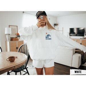 May include: White hoodie with a small graphic of a wave and a red sun. The hoodie has a drawstring hood and a front pocket. The person in the image is wearing a black baseball cap.