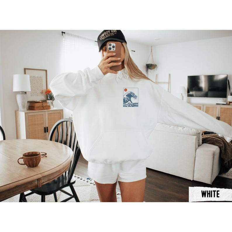 May include: White hoodie with a small graphic of a wave and a red sun. The hoodie has a drawstring hood and a front pocket. The person in the image is wearing a black baseball cap.
