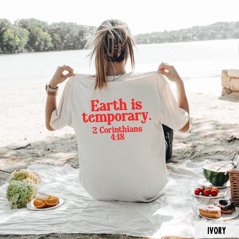 May include: Ivory t-shirt with red text that reads 'Earth is temporary. 2 Corinthians 4:18.' The person is sitting on a white blanket with food and drinks, including watermelon, tomatoes, and donuts. The background includes a lake and trees.