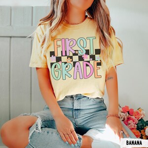K&ouml;nnte beinhalten: Ein gelbes T-Shirt mit den Worten "FIRST GRADE" in bunten Buchstaben und einem karierten Muster. Das T-Shirt hat einen Rundhalsausschnitt und kurze &Auml;rmel. Der Text ist in einer Retro-Schriftart mit floralen Akzenten gehalten. Das T-Shirt wird mit einer blauen Jeans getragen.