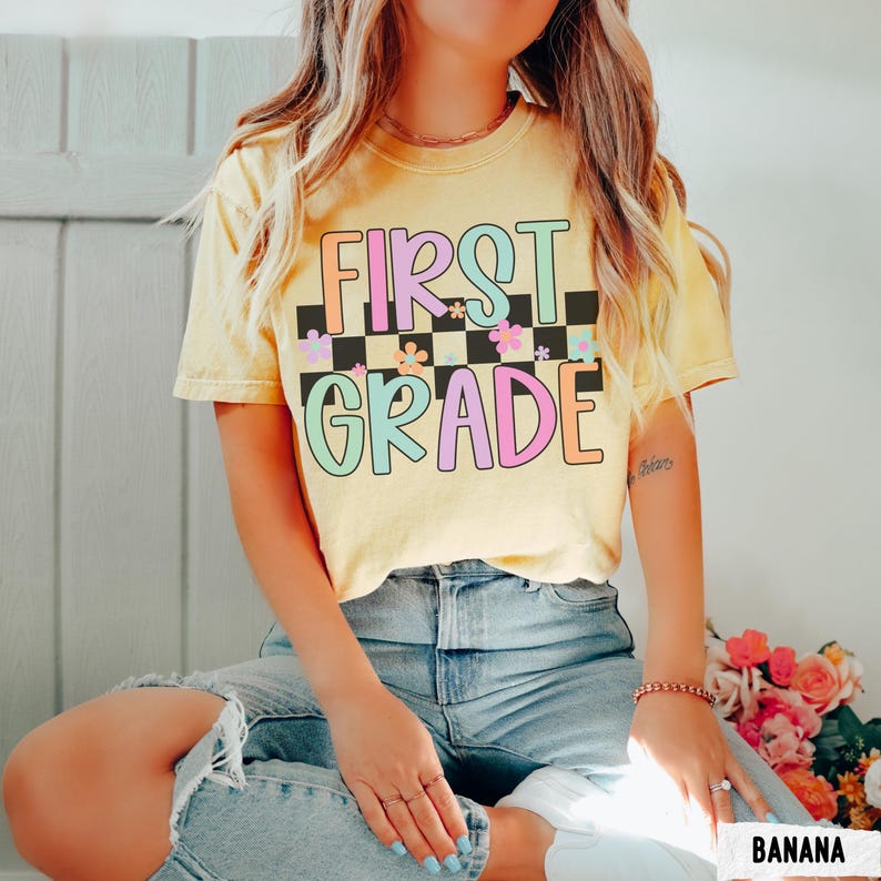 K&ouml;nnte beinhalten: Ein gelbes T-Shirt mit den Worten "FIRST GRADE" in bunten Buchstaben und einem karierten Muster. Das T-Shirt hat einen Rundhalsausschnitt und kurze &Auml;rmel. Der Text ist in einer Retro-Schriftart mit floralen Akzenten gehalten. Das T-Shirt wird mit einer blauen Jeans getragen.