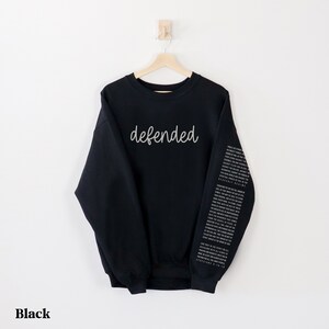 May include: Black sweatshirt with the word "defended" in a cursive font across the chest. The right sleeve features a vertical column of text. The word "Black" is at the bottom of the image.