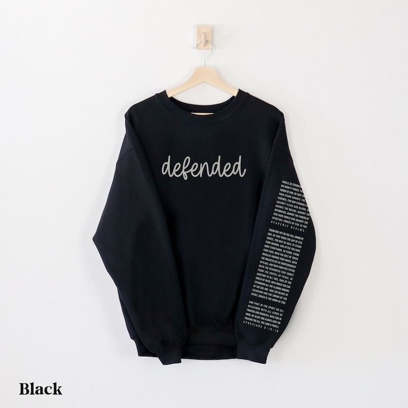 May include: Black sweatshirt with the word "defended" in a cursive font across the chest. The right sleeve features a vertical column of text. The word "Black" is at the bottom of the image.