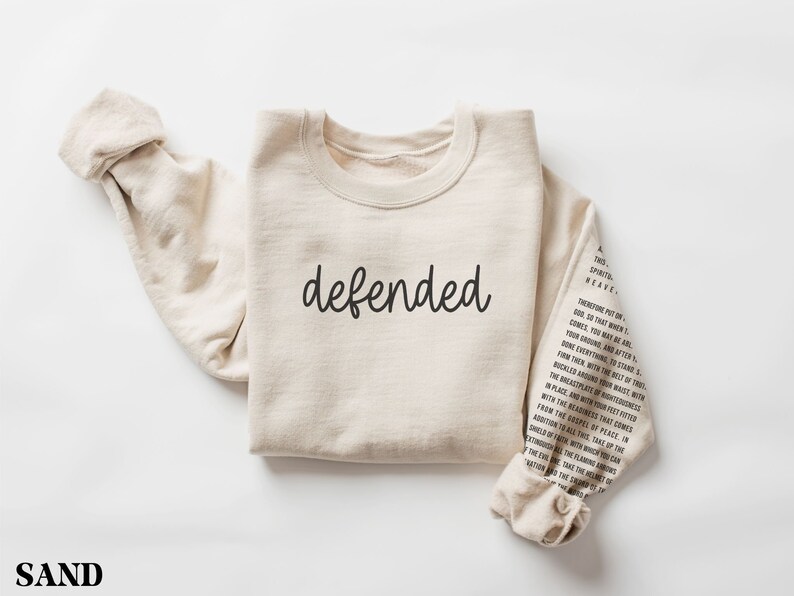May include: A sand-colored sweatshirt with the word "defended" in black script across the chest. The right sleeve features printed text. The word "SAND" is printed at the bottom.