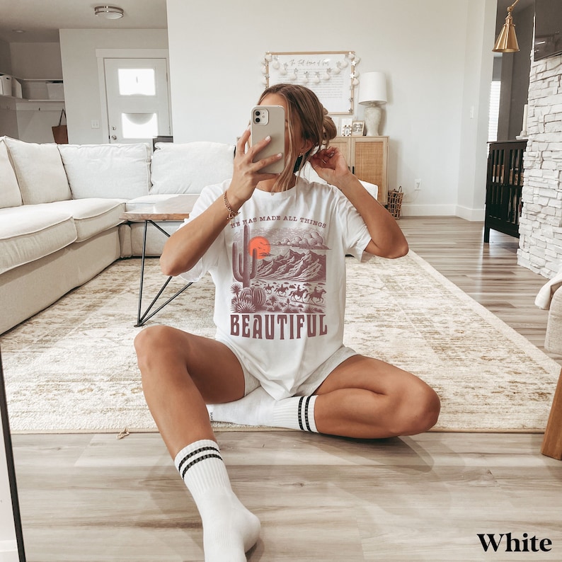 K&ouml;nnte beinhalten: Wei&szlig;es T-Shirt mit einem grafischen Druck einer W&uuml;stenlandschaft mit einem Kaktus, Bergen und einer untergehenden Sonne. Der Text "He has made all things beautiful" ist auf das Shirt gedruckt.