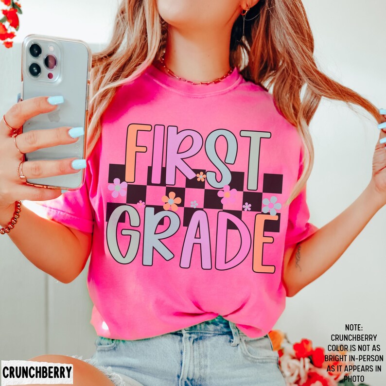 K&ouml;nnte beinhalten: Ein leuchtend pinkfarbenes T-Shirt mit dem Schriftzug "FIRST GRADE" in einem bunten Retro-Schriftstil. Das Design umfasst ein Schachbrettmuster und florale Akzente. Ein fr&ouml;hliches und farbenfrohes Kleidungsst&uuml;ck.