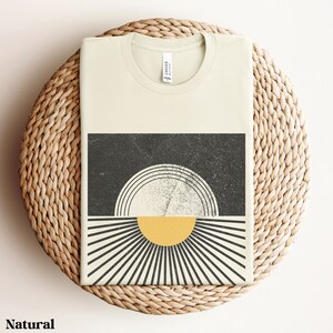 May include: A beige t-shirt with a graphic design of a black and white sun with a yellow center. The sun is surrounded by black and white rays.