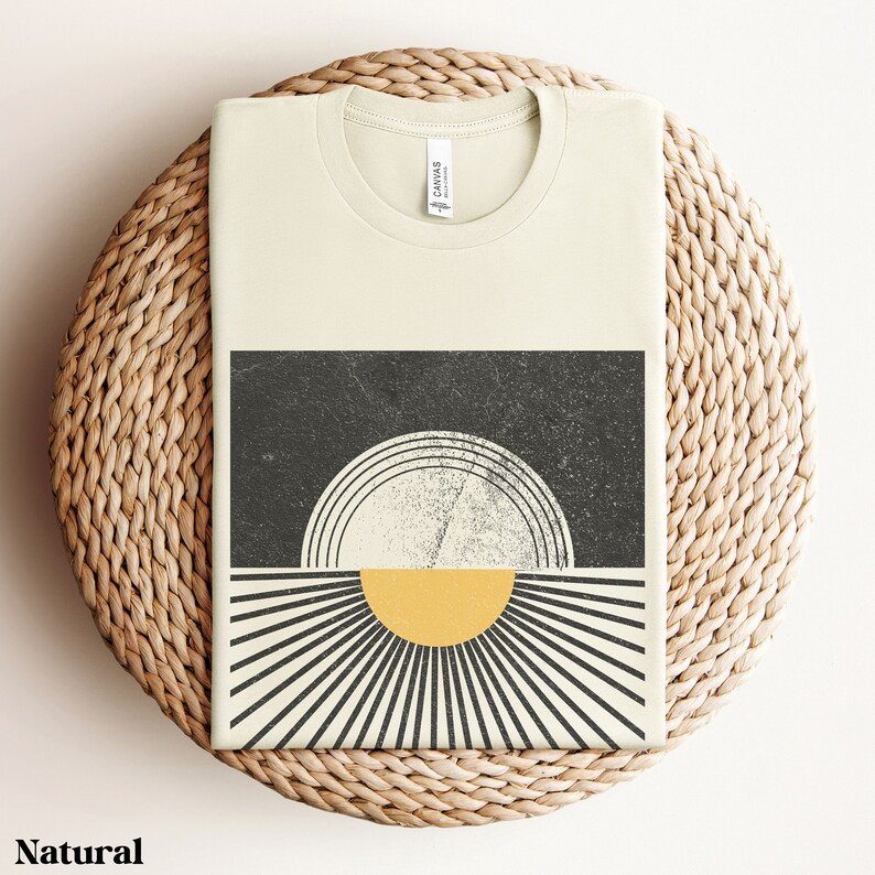 May include: A beige t-shirt with a graphic design of a black and white sun with a yellow center. The sun is surrounded by black and white rays.