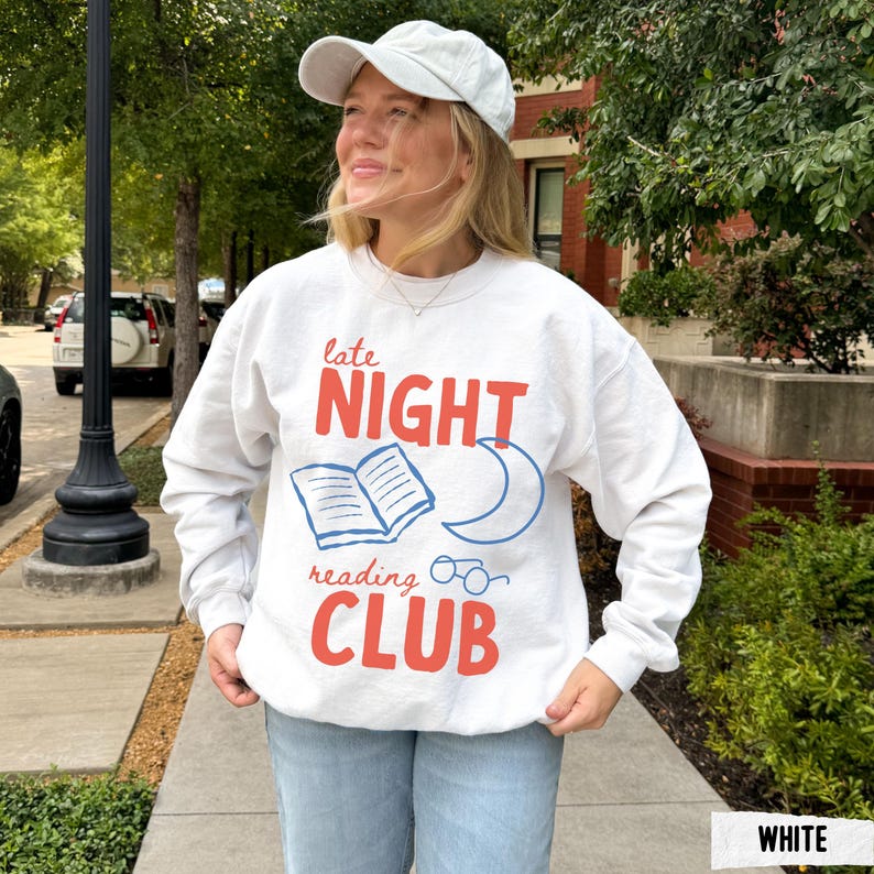 May include: White sweatshirt with the text "late NIGHT reading CLUB" in red and blue. The design includes an open book, a crescent moon, and glasses. The person is wearing a white cap and light blue jeans.