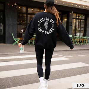 Pu&ograve; includere: Felpa nera con la scritta "STILL IN MY EMO PHASE" in un design circolare bianco sul retro. Il design include fulmini e un motivo a scacchiera. La persona indossa leggings neri, calzini bianchi e scarpe da ginnastica.