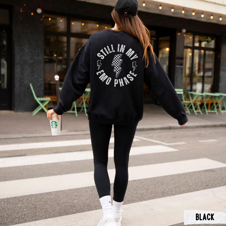 Pu&ograve; includere: Felpa nera con la scritta "STILL IN MY EMO PHASE" in un design circolare bianco sul retro. Il design include fulmini e un motivo a scacchiera. La persona indossa leggings neri, calzini bianchi e scarpe da ginnastica.