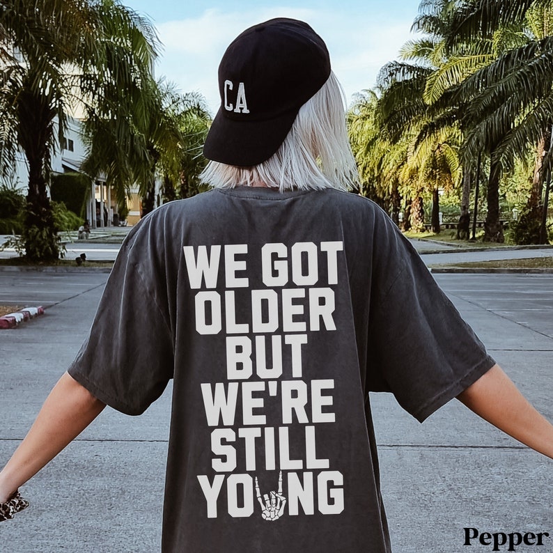 May include: Dark gray t-shirt with the text "WE GOT OLDER BUT WE'RE STILL YOUNG" in white block letters. A black baseball cap with "CA" in white is worn. The shirt features a skeleton hand making a rock and roll gesture.