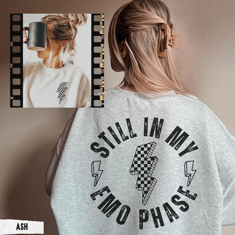 May include: Ash-colored sweatshirt with the text "STILL IN MY EMO PHASE" in a circular design, featuring a checkered lightning bolt graphic. The sweatshirt is a light gray color.
